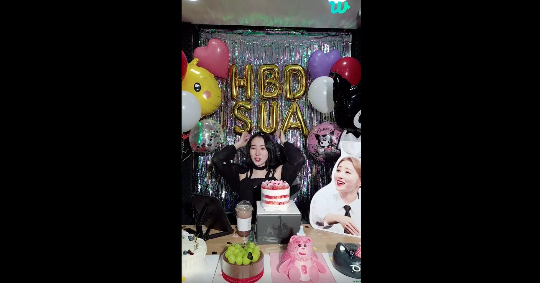 SuA cheers for her birthday with decorations and streamers in the background and cakes and gifts in the foreground