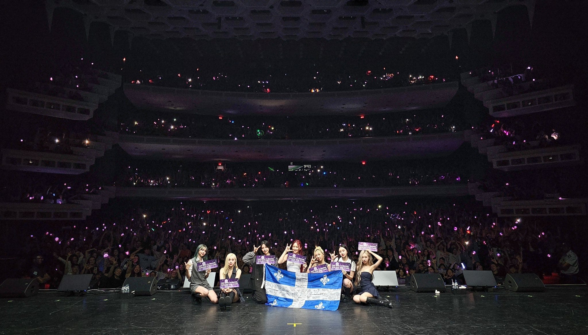 Dreamcatcher posing with the blue and white Quebec flag in front of their Montreal audience who have their lightsticks turned on