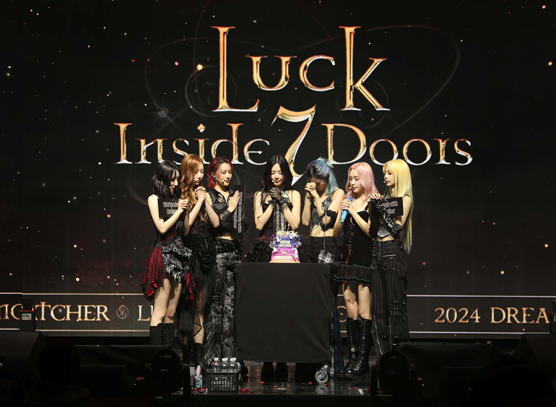 Dreamcatcher on-stage for their 7th anniversary content clasping their hands in prayer with a cake on stage.