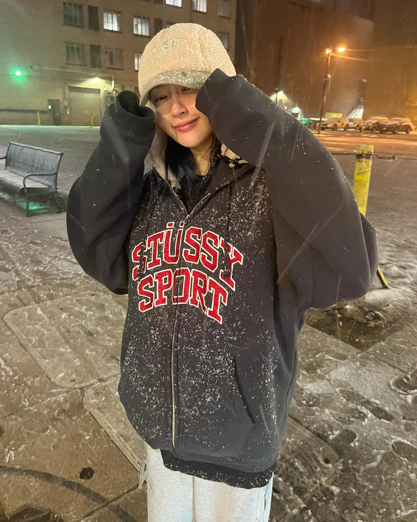 Siyeon, lightly covered with snow in a black hoodie reading STUSSY SPORT, smiling for the camera.