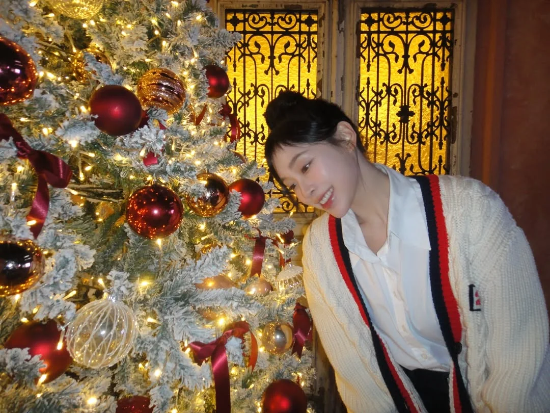 Gahyun in a white sweater smiling at a Christmas tree with red ornaments.