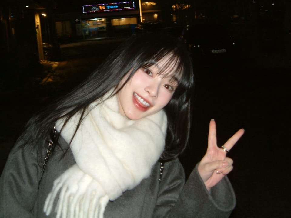 Gahyun dressed in a white scarf and grey coat smiling while giving the victory sign to the camera.