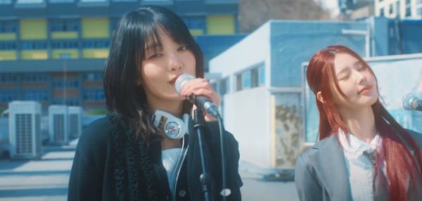 Minnie and Miyeon from (G)-IDLE, dressed in schoolwear, sing "Fate" over live microphones.