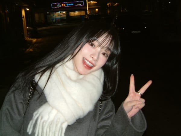 Gahyun dressed in a white scarf and grey coat smiling while giving the victory sign to the camera.